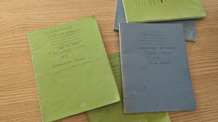A stack of weather observation notebooks on a wooden table. The top green notebook is labeled "Weather Observations, January 1999 to February 2000." A blue notebook beneath it is labeled "Weather Records, June 2002 to June 2003.