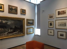 Cooper Gallery artwork near the lift. A large picture of a ship on a stormy sea and smaller pictures less distinct. A seat with orange cushions in the middle.