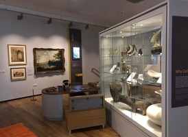 Cooper Gallery exhibits near the lift  including artworks, ceramics and pottery in a glass case. A wooden chair and table with children's activities such as piecing together a pot is in the middle of the room.