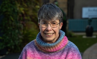 An older adult with short gray hair and glasses smiles at the camera. They are wearing a colorful, striped sweater with shades of purple, pink, and blue. The background is blurred and features greenery and buildings.
