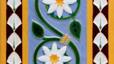 A decorative tile with two white flowers featuring yellow centers on a green vine. The background is blue with vertical yellow and brown patterned borders of white geometric shapes.