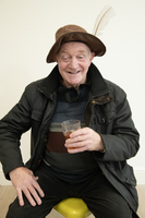 Older man in a feathered brown hat and jacket sits on a yellow stool, smiling and holding a drink in his right hand against a plain light-colored background.