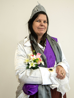 A woman in a silver crown, white and purple costume, and gray scarf holds a bouquet of artificial flowers, standing against a plain background.
