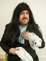 A man with a fake long black wig wears a suit and lace cuffs, holding a white cloth and posing humorously against a plain background.