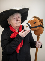 Elderly woman in a black hat and red scarf holds a toy stick horse and gestures toward its face, standing against a plain background.