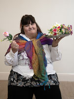 A woman with dark hair wears a white shirt, colorful scarf, and floral pants, holding a pink rose in one hand and a tray of artificial flowers in the other, sitting against a plain background.