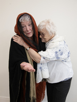 Two elderly women stand together; one in a dark shawl with a hood holds out her hand, while the other, in a white scarf, hugs her gently, both appearing emotional against a plain background.