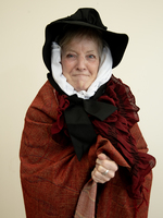 An older woman wearing a black bonnet, white scarf, and a red shawl stands in front of a plain background, smiling gently at the camera.