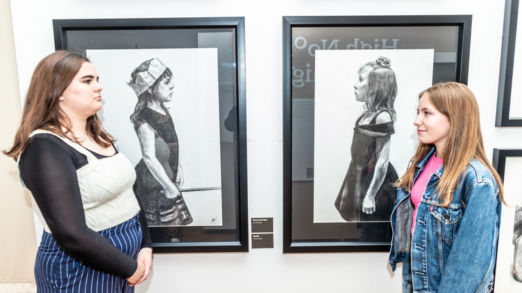 Two women stand in front of black-and-white portraits in an art gallery. The portraits feature young girls in various poses. The women are looking at the artworks and each other, seemingly engaged in conversation.