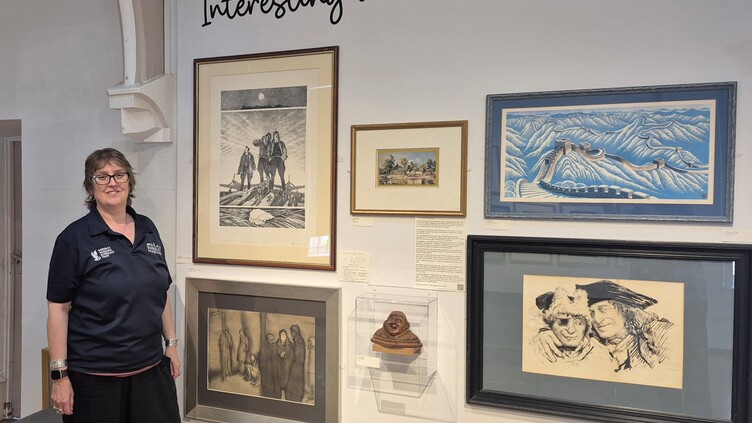 A woman in a navy shirt stands beside a wall displaying various framed artworks and a small sculpture. Above the art, text on the wall reads, “Interesting and unknown...”.