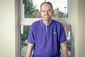 A man in a purple polo shirt sits by a window, looking at the camera. The background shows a view of trees and buildings outside.