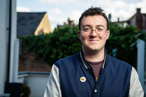 A person with glasses wearing a blue and gray jacket stands outside, smiling. They have a pin on their jacket. The background features greenery and blurred rooftops under a partly cloudy sky.