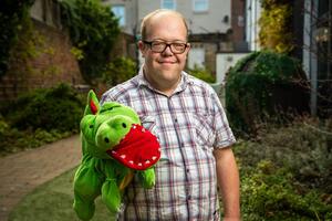 A person wearing glasses and a plaid shirt smiles while holding a green dragon puppet with a red mouth. The background features a brick wall, greenery, and a pathway.