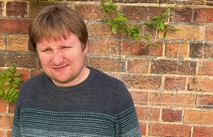 A person with short brown hair wearing a dark knit sweater stands in front of a brick wall with some greenery. The person is smiling and squinting slightly.