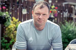A man in a gray Adidas shirt is sitting outdoors, looking at the camera. The background features a wooden fence decorated with string lights and hanging plants, with greenery and flowers visible around him.