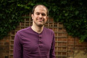 A man stands in front of a garden backdrop with ivy and trellis on a brick wall. He is wearing a purple long-sleeve shirt and is smiling slightly.
