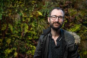 A person with glasses and a beard is standing outdoors against a backdrop of lush greenery, wearing a dark jacket with a fur-lined hood. They have a slight smile and their hair is short.